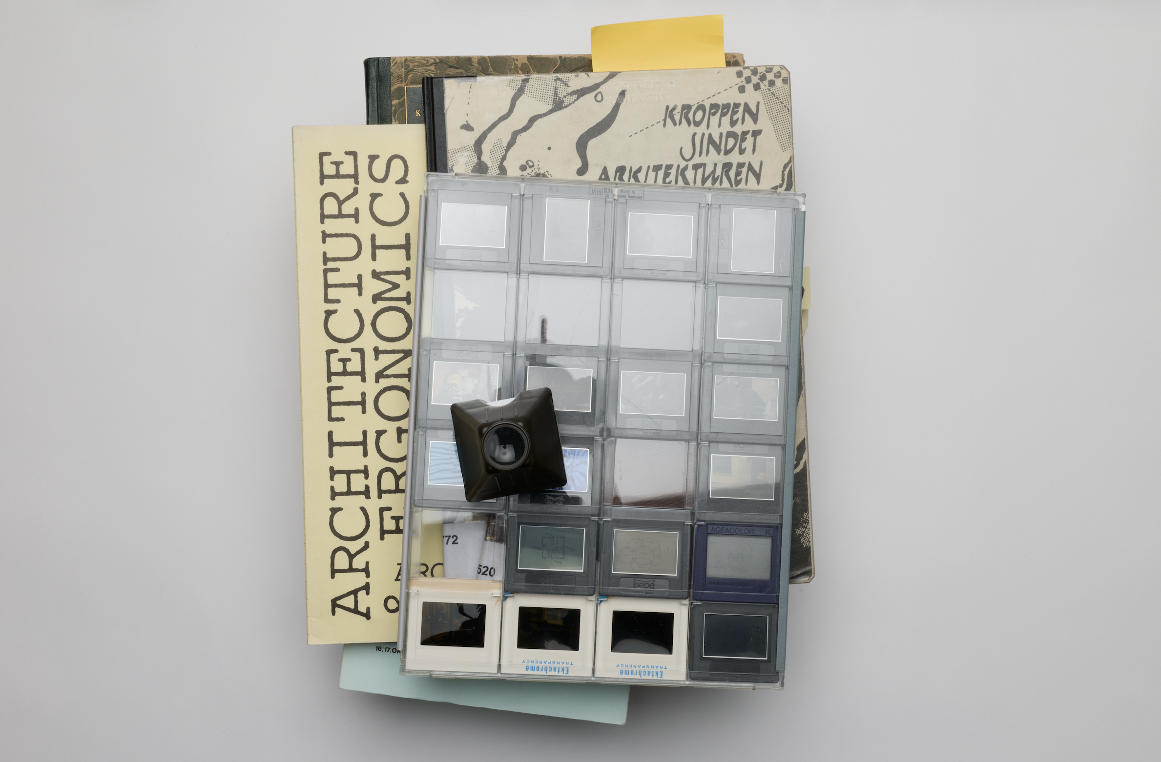 A stack of archival material rests on a light surface, topped with a clear sheet of photo slides arranged in a grid. A small black slide viewer rests on top, and sticky notes stick out from the pages. Photograph by Christine Bjerke