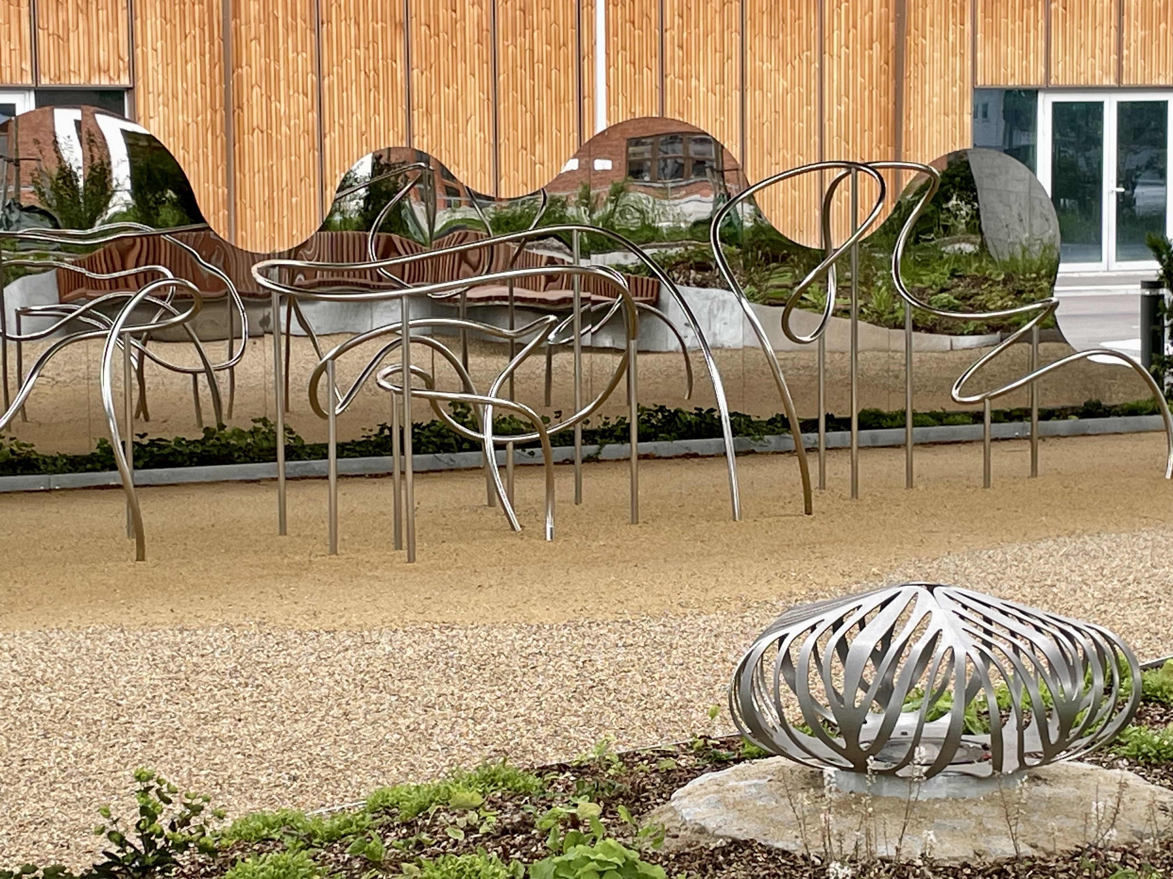 Public space with mirror in the background, sinewy metal apparatus for movement, gravel ground and a lantern in the front of the image