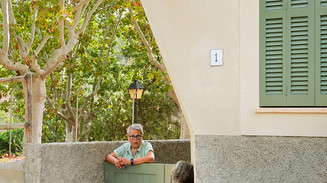 Interior with exposed stone walls and open rooms; two women talk by a green garden gate.