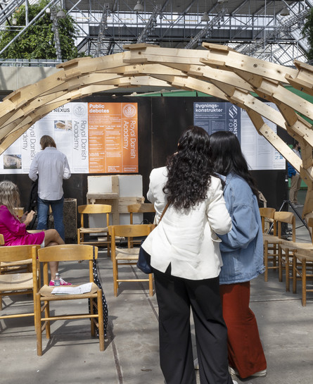 Wooden arch structure with chairs underneath, surrounded by exhibition displays and visitors.