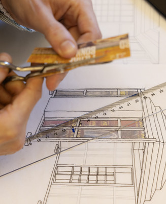 Hænder klipper materiale til en arkitekturmodel over en bygningstegning.