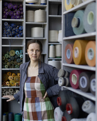 The image is portraying Trine in front of shelves filled with cones of yarn. She is wearing a blue jacket and a checkered dress in red, green, blue and white.