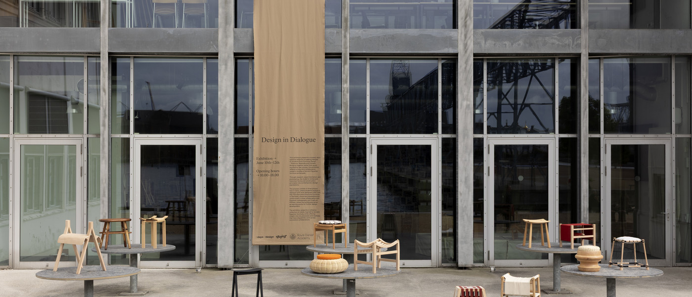 Design exhibition banner on a glass building façade with wooden stools displayed outside.