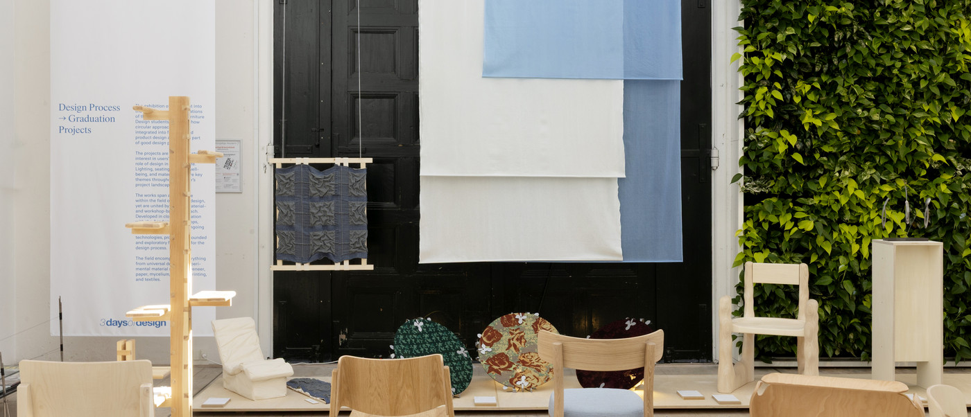 Exhibition space with wooden chairs, textiles, and wall text “Here & Now”