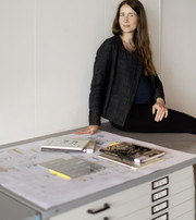 Color photograph of Christine Bjerke sitting on a archive furniture with archival materials next to her. 