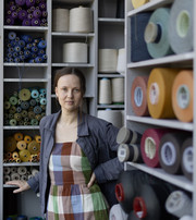 The image is portraying Trine in front of shelves filled with cones of yarn. She is wearing a blue jacket and a checkered dress in red, green, blue and white.