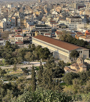 Long ancient colonnaded building surrounded by trees, with a dense cityscape behind.