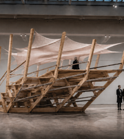 Visitors explore a large timber installation with stairs and stretched fabric at an exhibition.