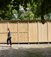 Woman walking past a wooden structure with smooth and woven panels beneath green trees.