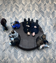 People are sitting and lying on a circular bench surrounded by herringbone-patterned wooden flooring.
