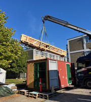 A crane lifts a long timber lattice structure above a red container next to an industrial building.