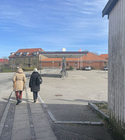Two people walk along the pavement past an open square with an electric vehicle charging point and red-brick buildings.