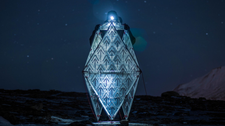 Person with headlamp inside a glowing, diamond-shaped shelter on rocky ground under a starry night sky.