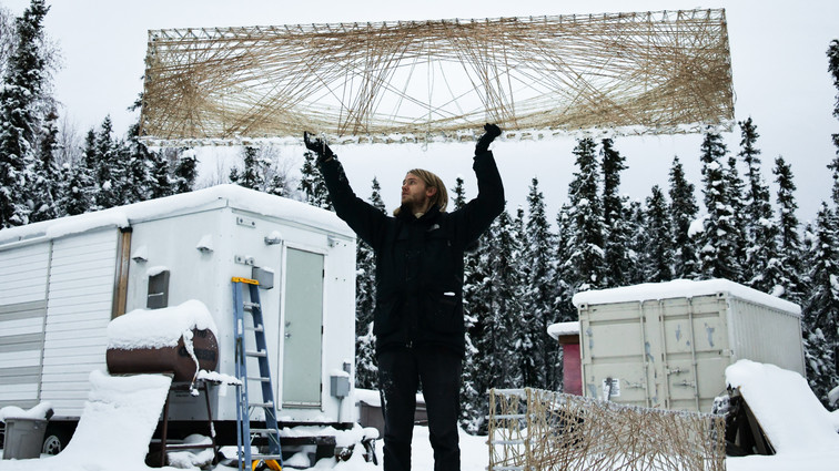 Person holding a large rectangular wooden lattice above their head in a snowy outdoor setting with cabins and trees.