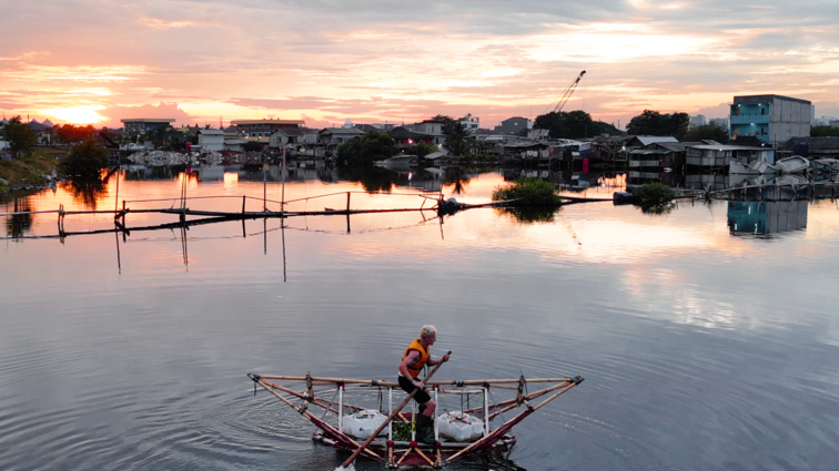 Person paddling a small triangular raft on calm water at sunset with houses and trees in the background.