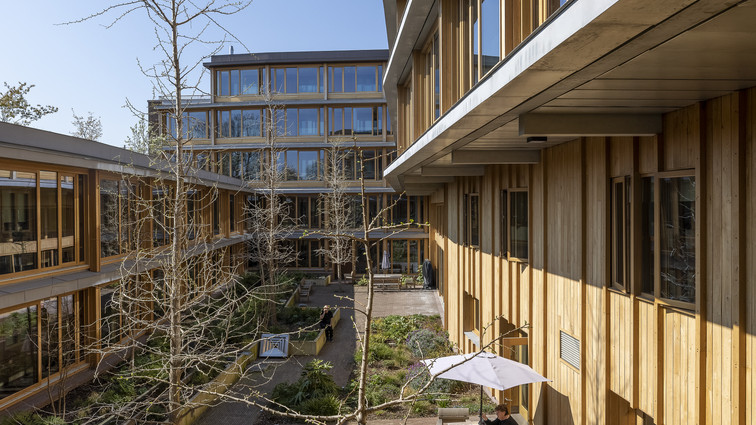  Gårdrum mellem moderne bygninger i træ med siddepladser og beplantning.