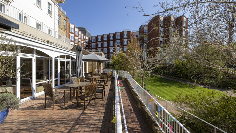  Terrasse med borde og stole langs bygning ved grønt gårdmiljø.