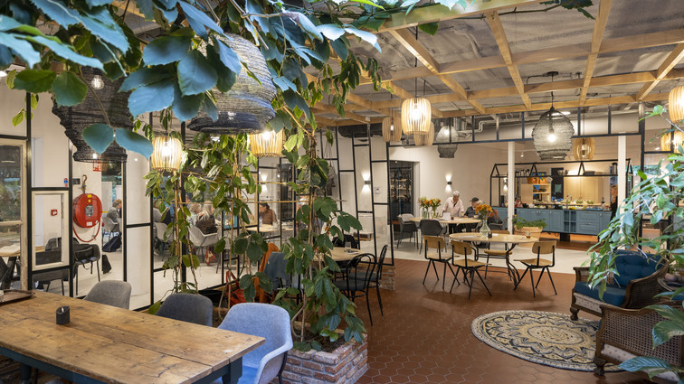Lyst fællesområde med caféborde, grønne planter og åben indretning.