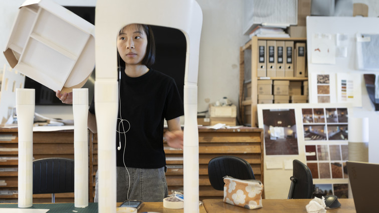 Person arbejder i værksted med en hvid stol/designprototype omgivet af modeller og materialer.