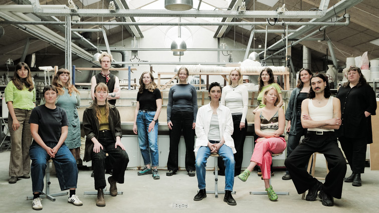 Group portrait of people seated and standing in a bright workshop with shelves, tools, and industrial lamps in the background