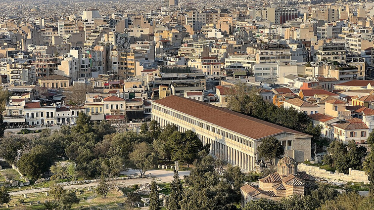Long ancient colonnaded building surrounded by trees, with a dense cityscape behind.