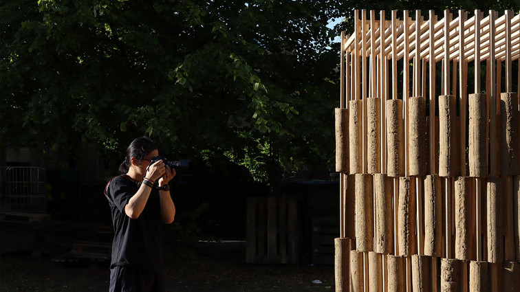 Photographer documenting a structure of timber slats and earth-toned elements.