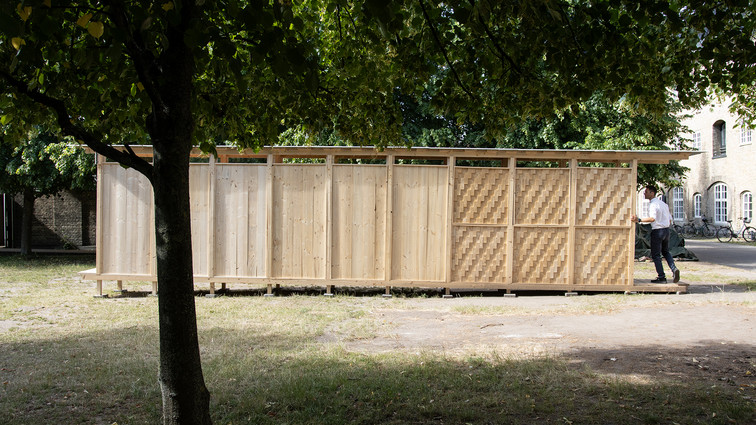 Long wooden pavilion with patterned wall panels beneath large trees.