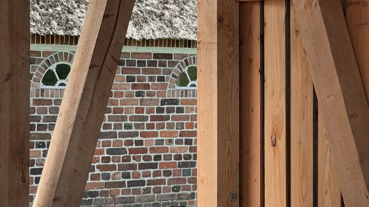 Indvendig trækonstruktion med skrå stolper og plankegulv foran en gammel murstensbygning.