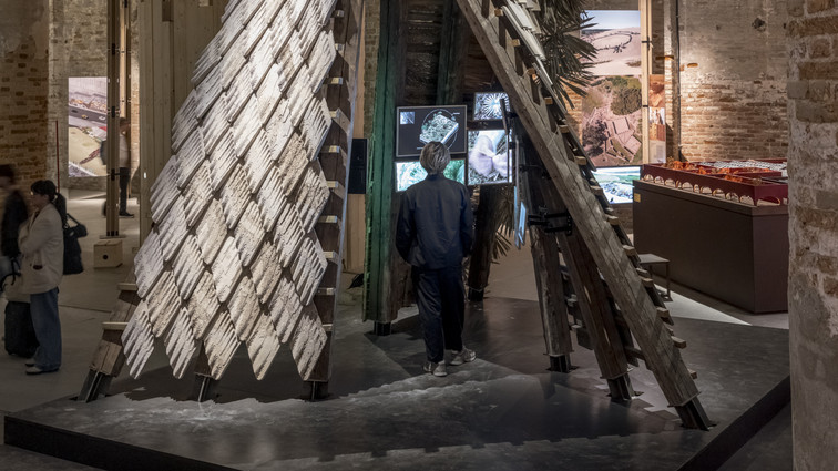 Large, tent-like installation of timber and overlapping panels in an exhibition hall with visitors.