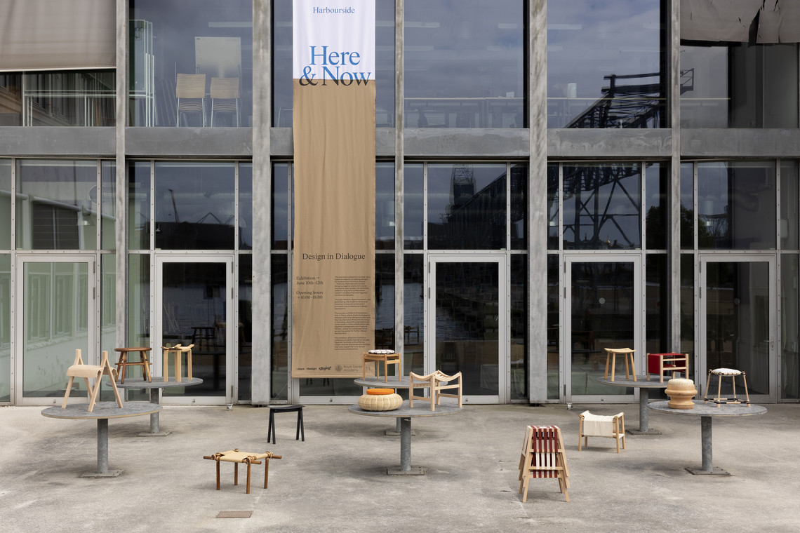 Design exhibition banner on a glass building façade with wooden stools displayed outside.
