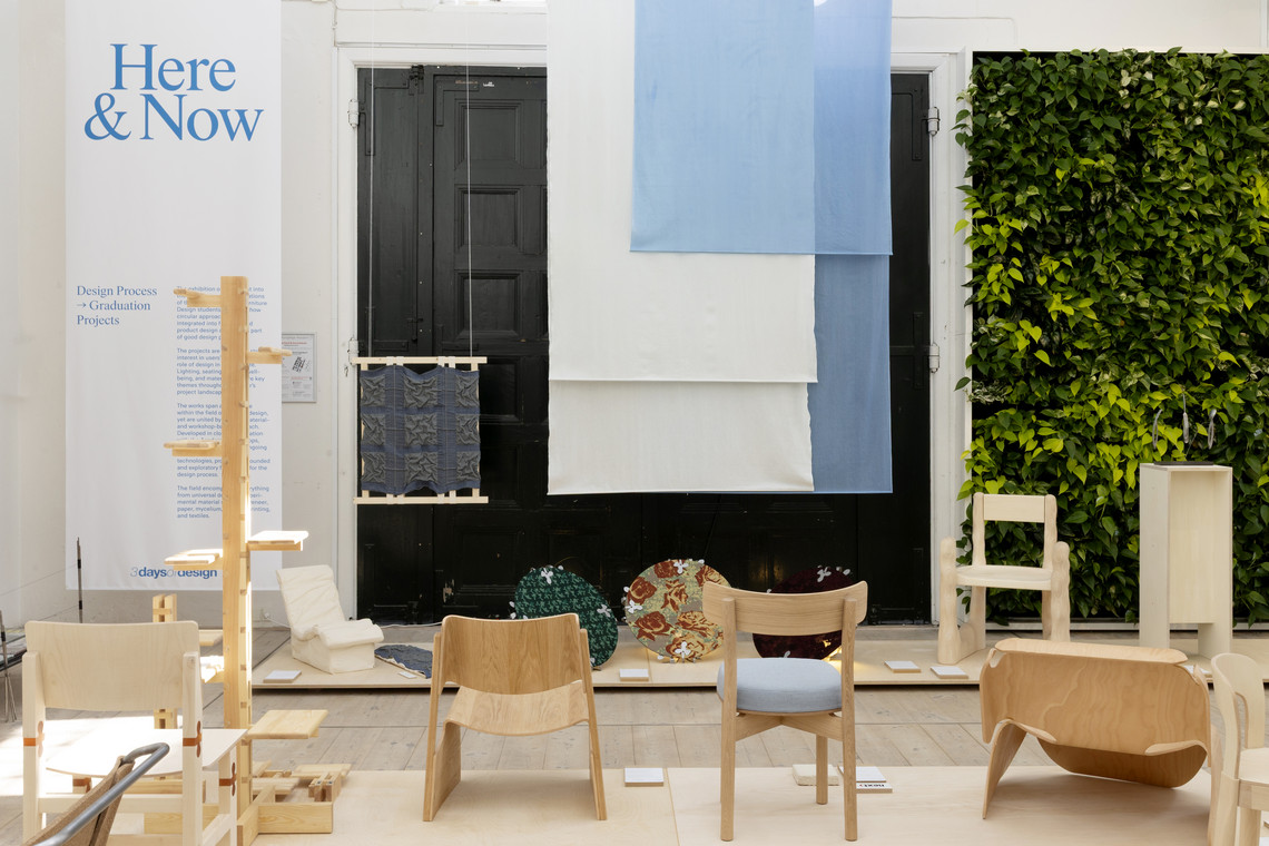 Exhibition space with wooden chairs, textiles, and wall text “Here & Now”