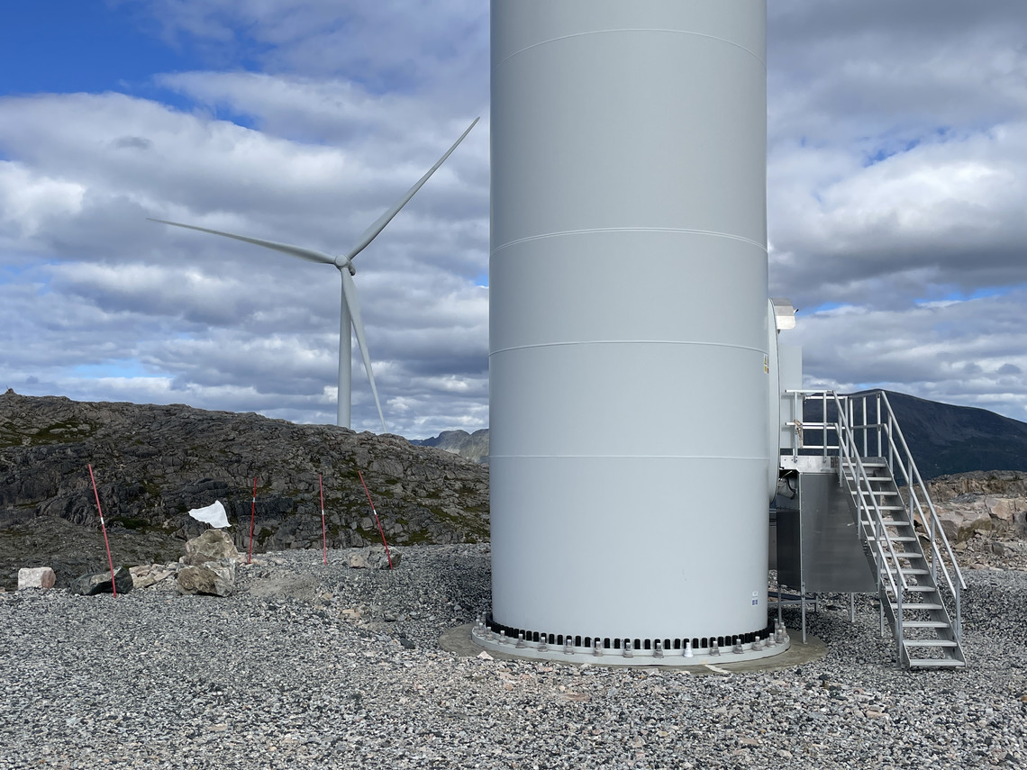 Color image of the base of a windmill with a staircase on a mountain platform with gravel in the foreground, and mountaintops and a windmill with three blades in the background. Blue sky with light clouds.