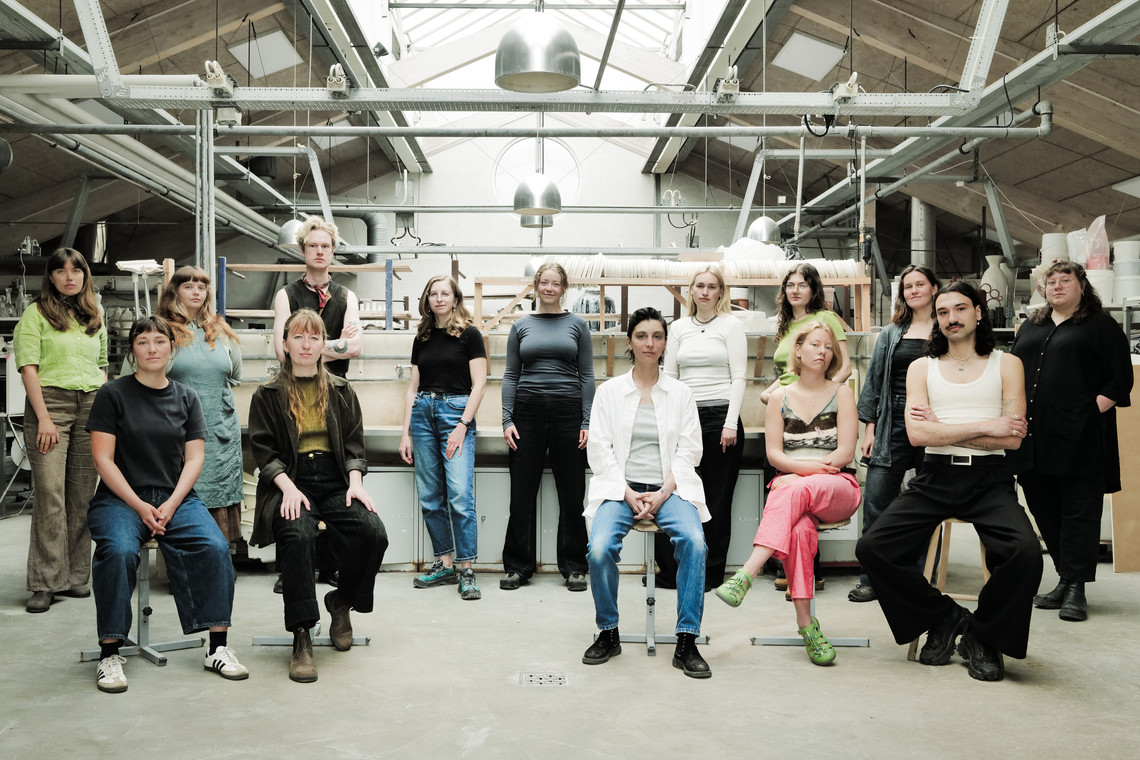 Group portrait of people seated and standing in a bright workshop with shelves, tools, and industrial lamps in the background