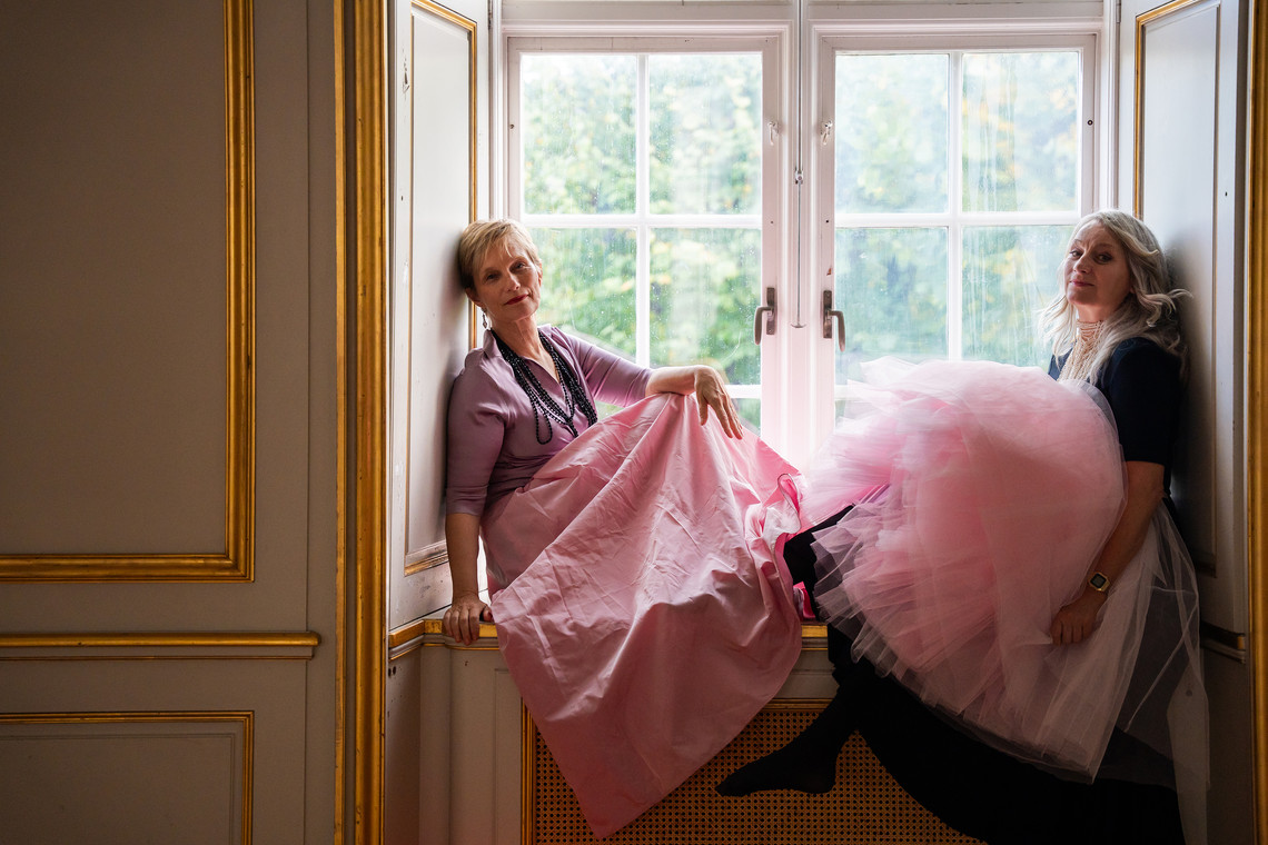 Two ladies sitting in a window. Both wear pink skirts.