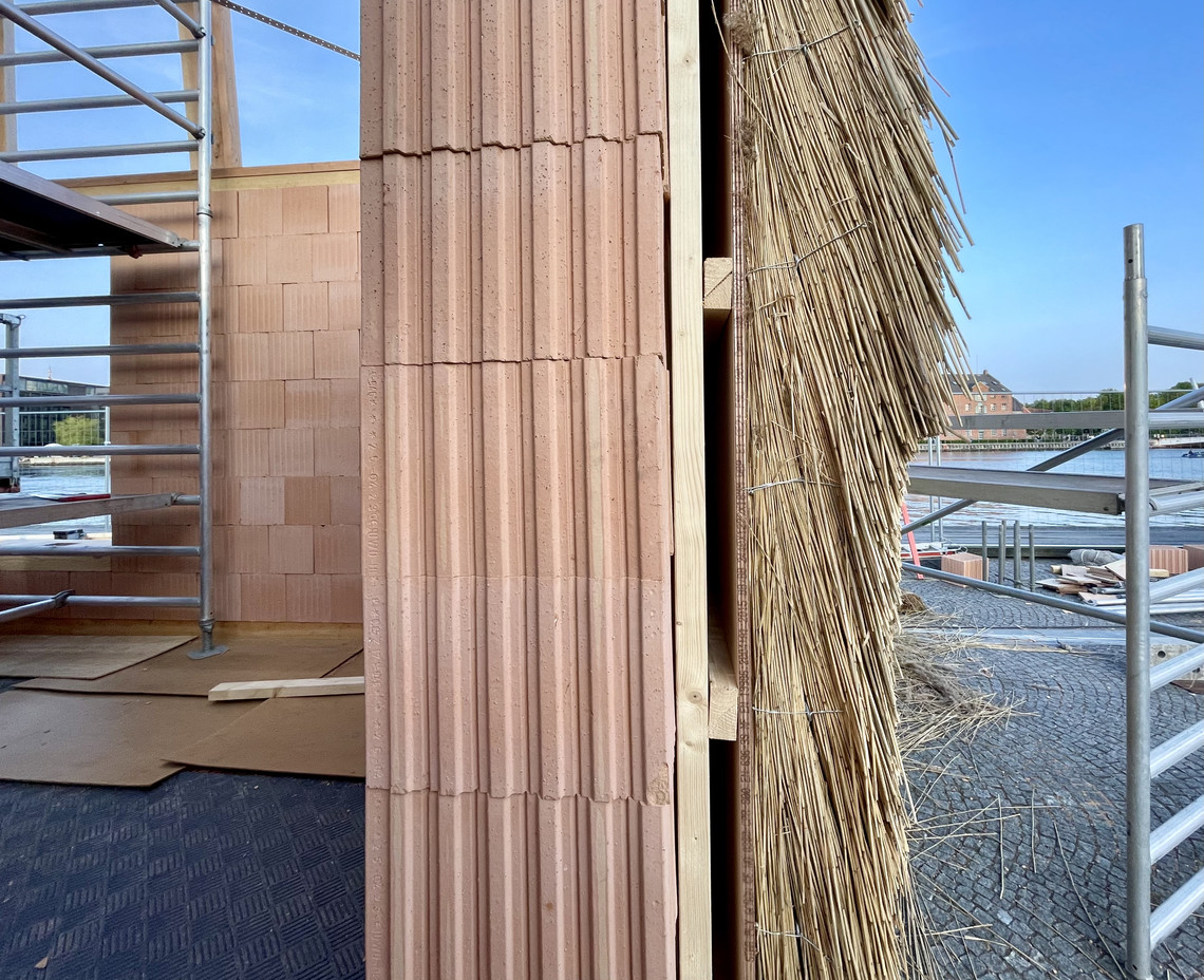 Close-up of a wall under construction with red clay blocks, timber framing and bundles of reeds.