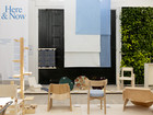 Exhibition space with wooden chairs, textiles, and wall text “Here & Now”