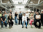 Group portrait of people seated and standing in a bright workshop with shelves, tools, and industrial lamps in the background