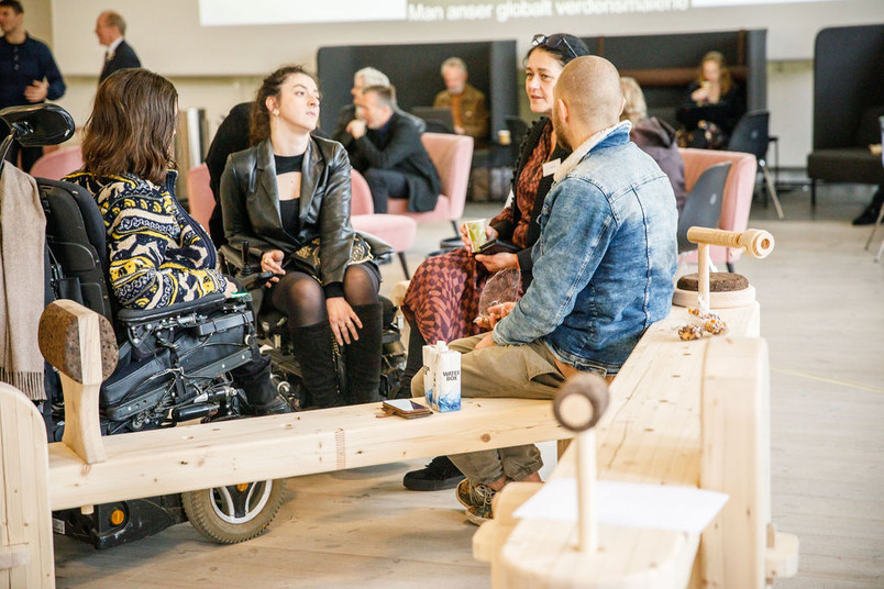 Group of people sitting and talking on wooden benches in an open meeting space with a screen in the background.