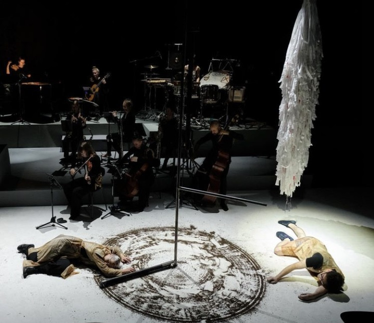 In the background, musicians on a stage; in the foreground, two actors lying on the floor