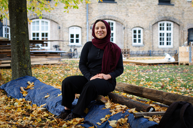 Young woman in black clothing and a burgundy scarf sitting on a tree trunk surrounded by fallen autumn leaves.