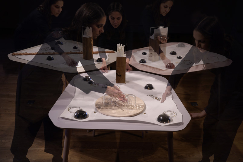 Art installation with hands interacting with objects on a geometric table in dim lighting.