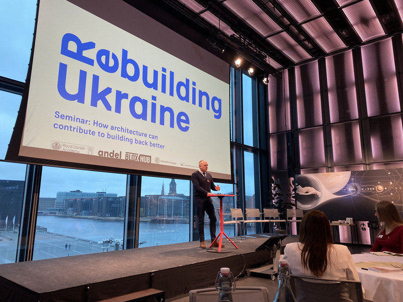 A speaker is standing on a stage in front of a large screen displaying the text ‘Rebuilding Ukraine’ during a seminar.