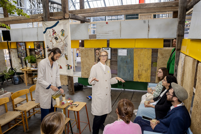 People in white lab coats are demonstrating something at a table in front of a seated audience inside a wooden structure.