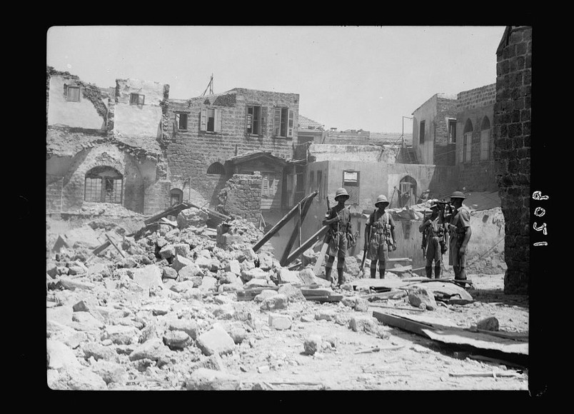 Uniformed individuals standing among the rubble of damaged stone buildings.