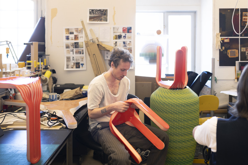 Person working in a workshop assembling a red, molded chair surrounded by design prototypes and materials.