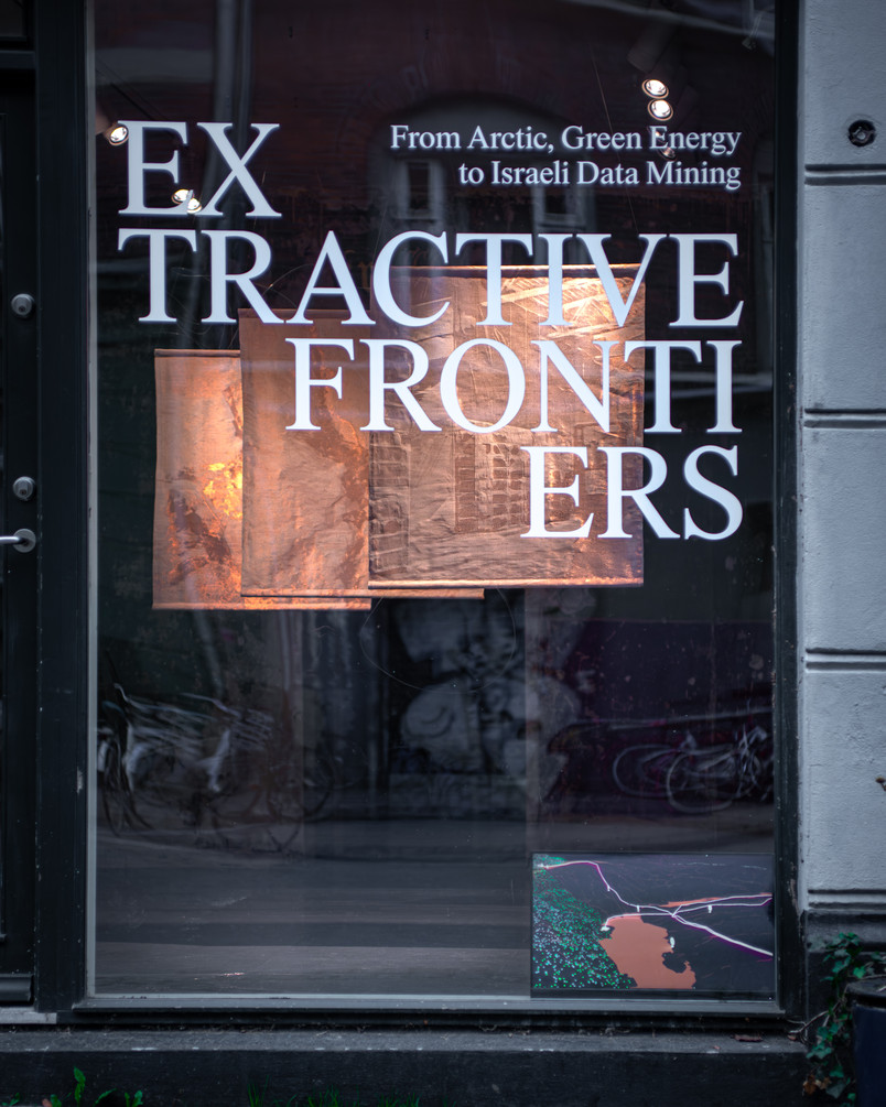 Storefront window with big letters that says EXTRACTIVE FRONTIERS across the window. Behind the window is a small screen with a colorful image in the lower right corner, and in the room there are three copper colored images hanging from the ceiling.
