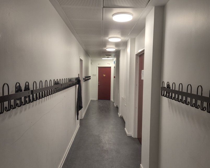 Photo of a narrow hallway in the school. Floors are linoleum, walls are white. There are no windows, a repetition of doors along the hall and coathangers on both sides