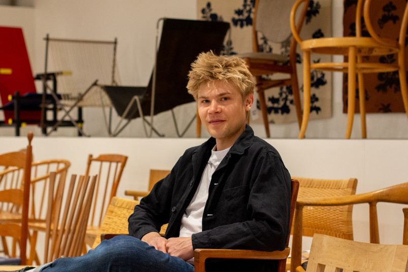 Person seated among wooden chairs in a design exhibition