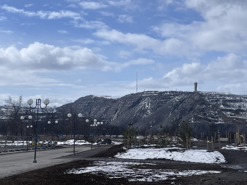 Landscape image of a mountain in the background and an open area with asphalted road with streetlights in the front. Blue sky dotted with clouds. The mountain and flat area is dark and barren with remains of snow