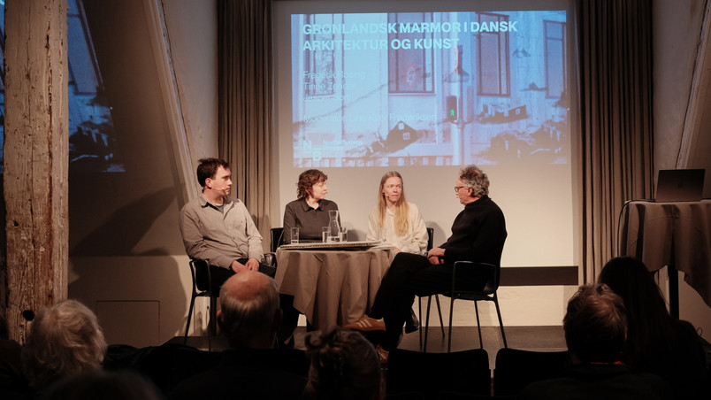 Image of four people in conversation sitting around a table on a stage, with a background image from the artist film "Honeycomb Image/Archive Cladding" by Eva La Cour and Tinne Zenner. Text on the image says "Greenland Marble in Danish Architecture and Art - Frederik Rosing, Tinne Zenner, Jesper Sort. Moderator: Line Kjær Frederiksen. Nordatlantens Brygge."